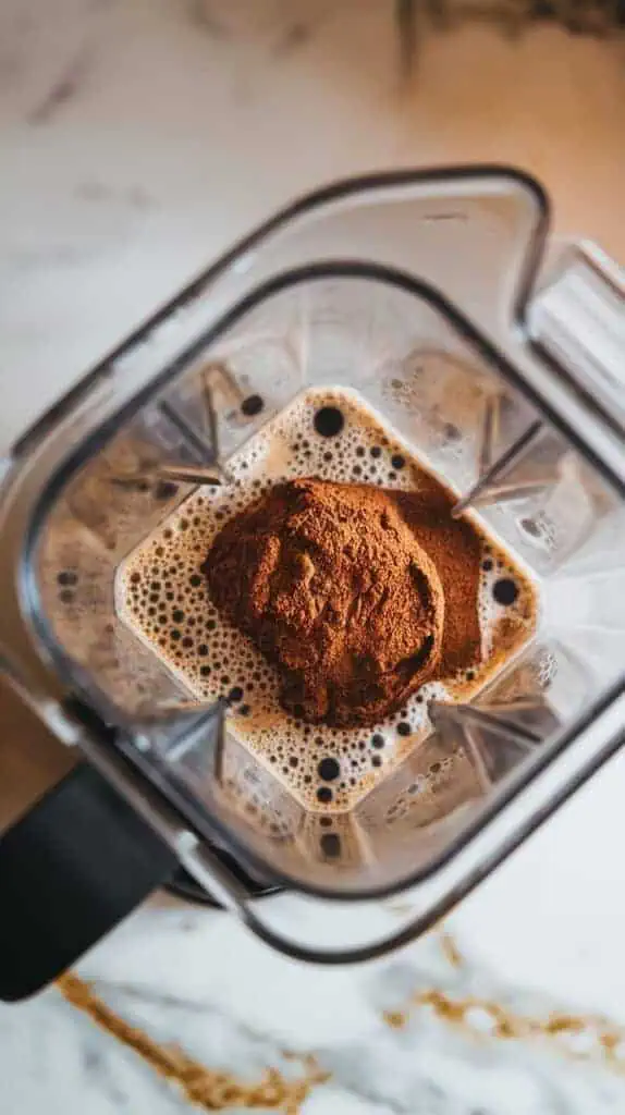 A blender filled with coffee, milk, and a scoop of cookies and cream protein powder, mid-blend, with frothy bubbles starting to form