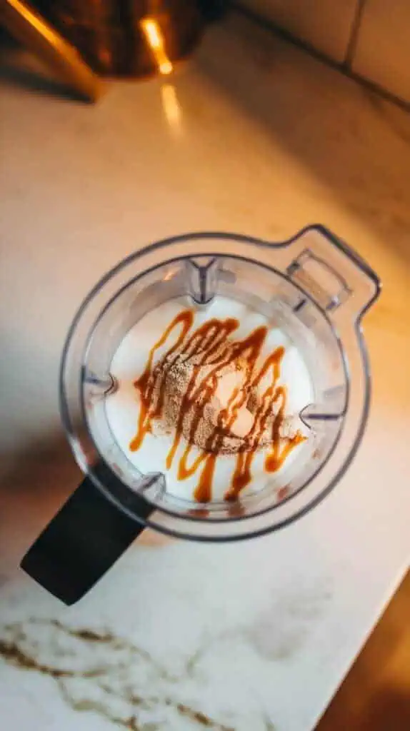 A blender cup filled with almond milk, vanilla protein powder, a drizzle of caramel sauce, and vanilla extract, frothing into a creamy mixture