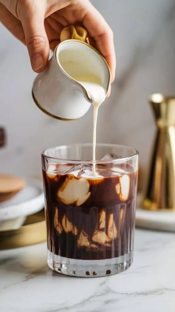 A hand pouring heavy cream from a small pitcher into a rocks glass filled with chocolate syrup, vodka, and coffee liqueur, creating swirls in the drink. 