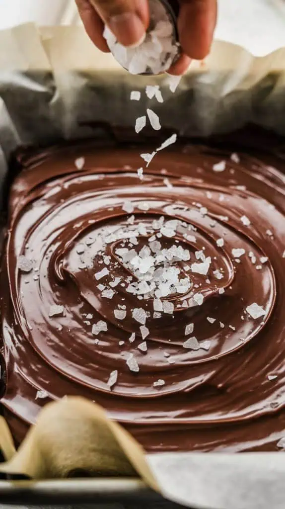 A super close-up shot of flaky sea salt being sprinkled onto the glossy surface of the fudge