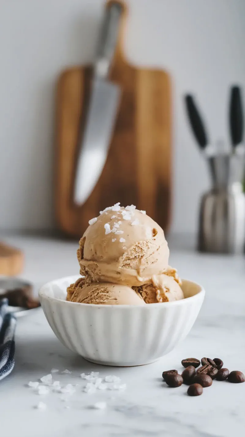 A bowl of salted caramel coffee ice cream topped with sea salt, surrounded by coffee beans.