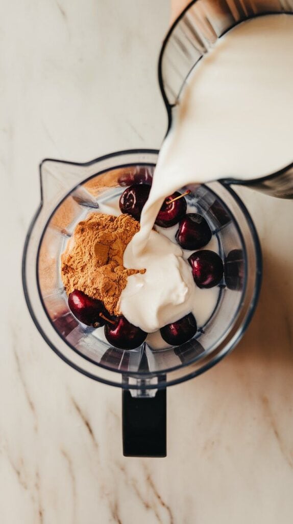 almond milk being poured into blender with protein powder and cherries, Greek yogurt dolloped on top