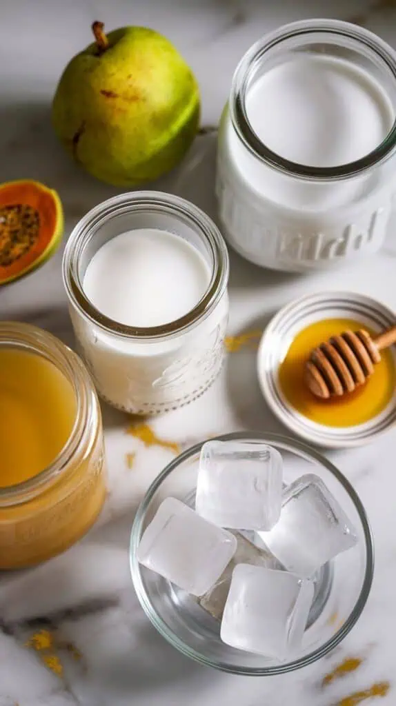 ingredients for guava coconut milk tea: guava juice in a clear glass jar, coconut milk in a second clear glass jar, cooled green tea in a third clear glass jar, honey on a small white plate, and clear ice cubes in a shallow clear dish