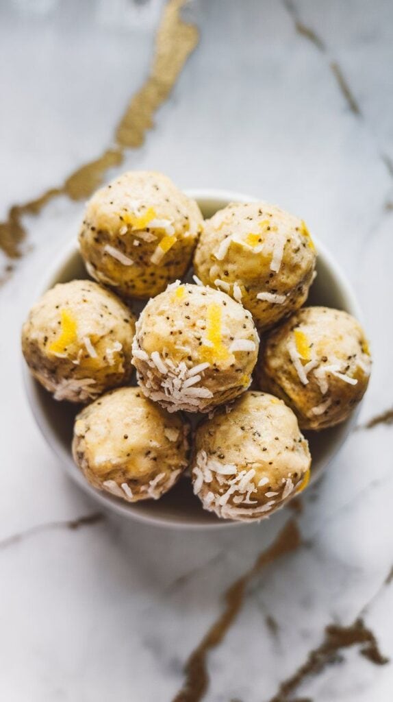 lemon poppy seed protein balls arranged in a small white dish, dusted lightly with shredded coconut, with flecks of poppy seeds and lemon zest visible.