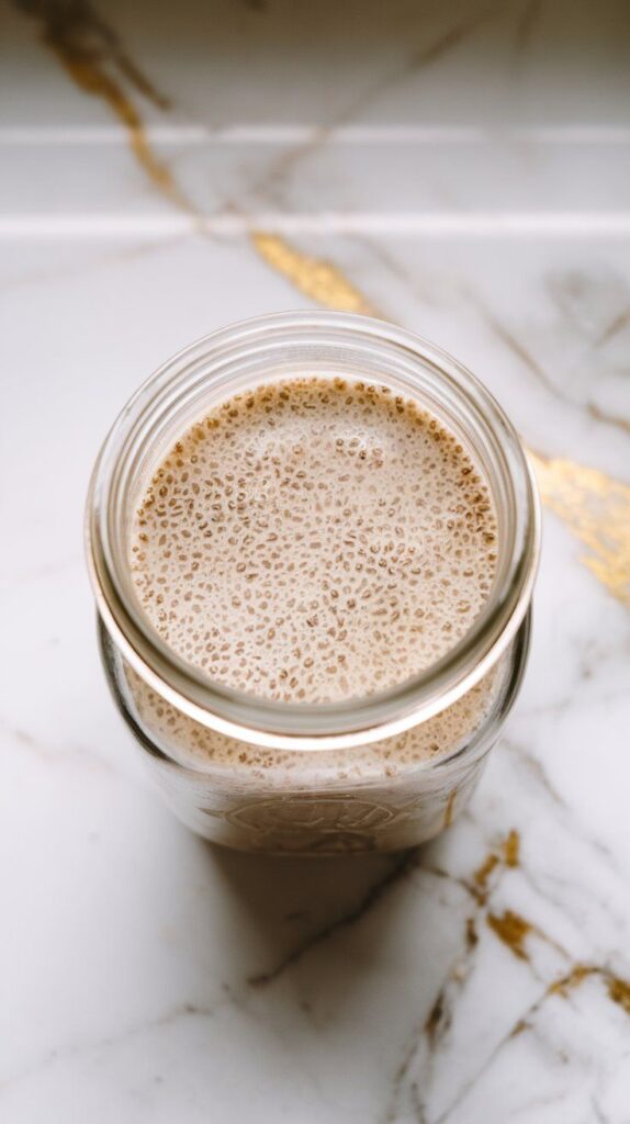 unsweetened almond milk, dry chia seeds, maple syrup, and vanilla extract stirred together into a pale, speckled liquid in a clear glass jar, texture just starting to thicken