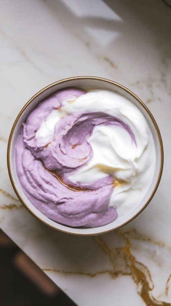 thick Greek yogurt and mashed ube being swirled together in a bowl, creating a marbled purple and white pattern with visible streaks of honey and vanilla