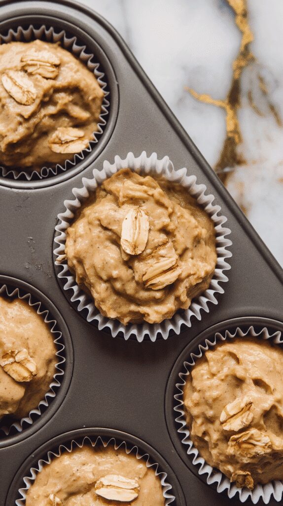  muffin batter scooped into paper liners inside a muffin tin, batter slightly lumpy with visible oats, on a white marble counter with hints of gold