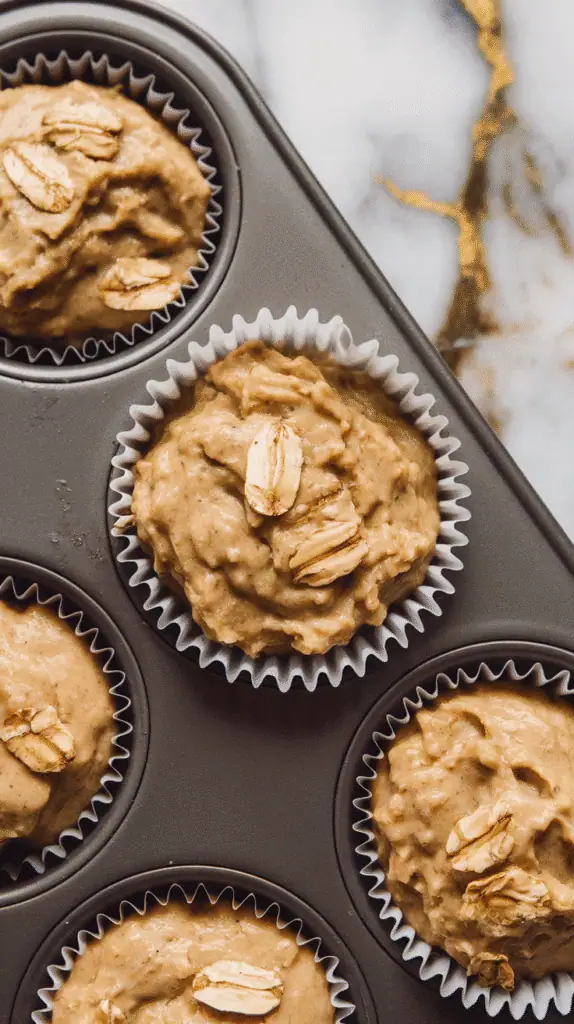  muffin batter scooped into paper liners inside a muffin tin, batter slightly lumpy with visible oats, on a white marble counter with hints of gold